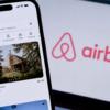 Airbnb app displayed on a smartphone with the company logo visible on a screen behind it.