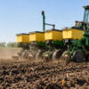 A green and yellow John Deere tractor pulls a multi-row seed planter across a tilled, soil-covered field